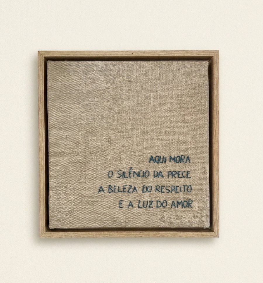 Frase 'Aqui mora o silêncio da prece, a beleza do respeito e a luz do amor' bordada à mão por Nara Lombardi em tela de linho com chassi de madeira